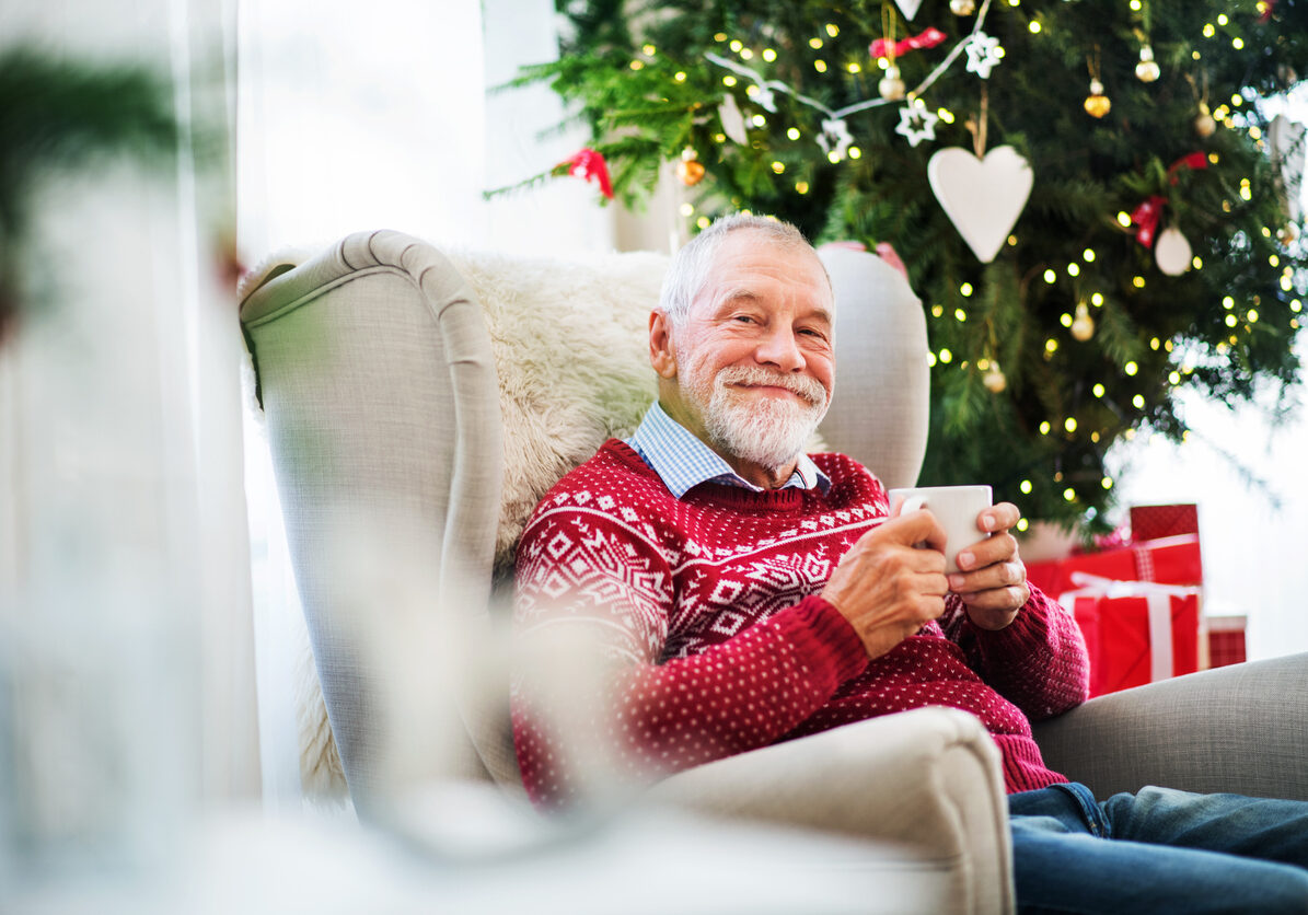 A portrait of senior man sitting on armchair at home at Christmas time, holding a white cup.