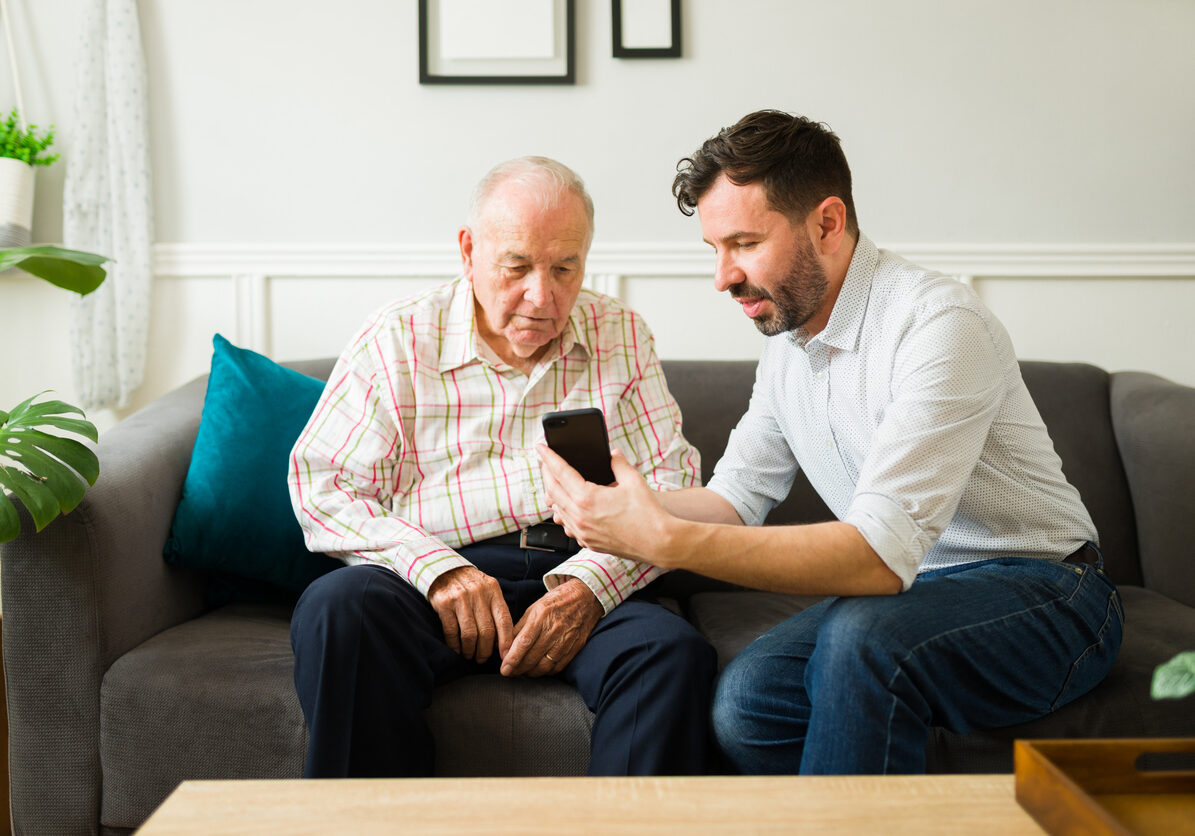 Happy caucasian son teaching his old man to text on the smartphone and helping to make calls to family