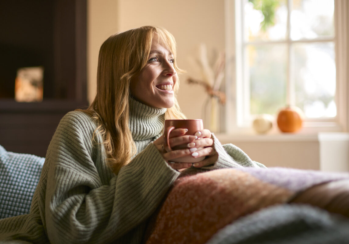 Smiling Woman At Home Wearing Winter Jumper With Warming Hot Drink Of Tea Or Coffee In Cup Or Mug Woman heating home on a budget
