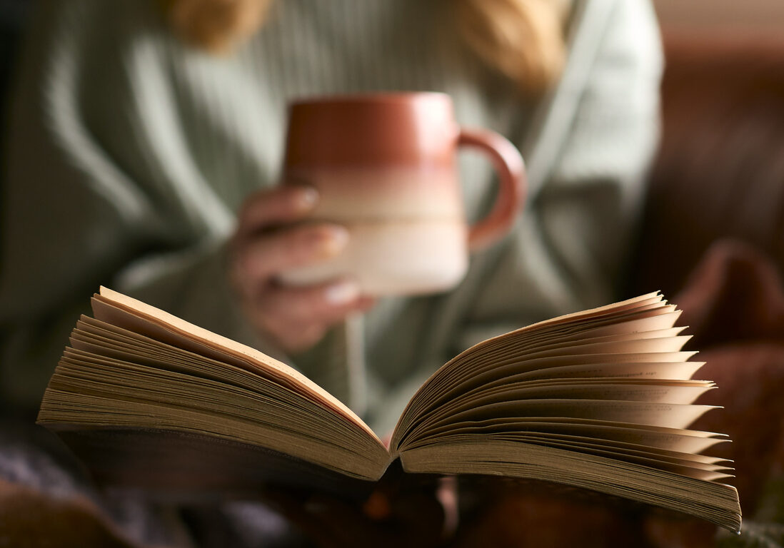 Close Up Of Woman At Home In Winter Jumper With Warming Hot Drink In Mug Reading Book