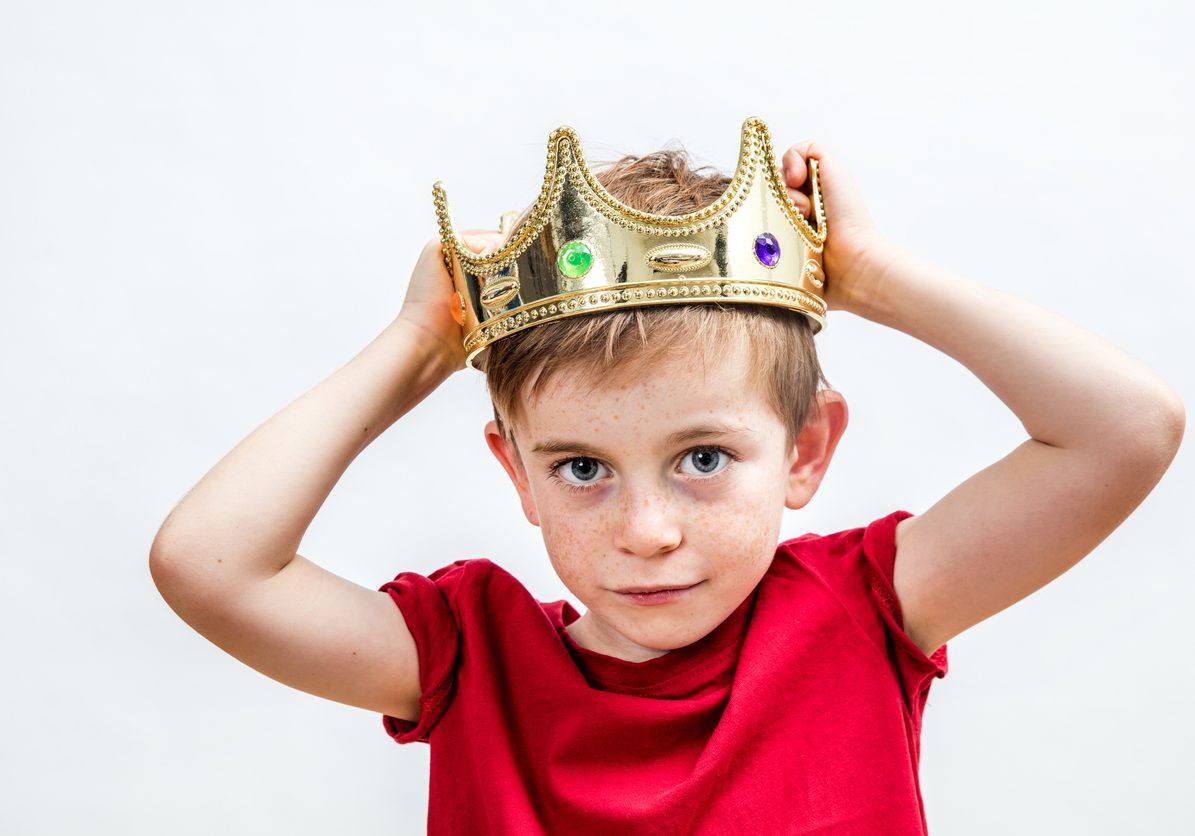 happy education and childhood concepts with an adorable 7-year old boy holding a golden king crown on his head as a wise spoiled child, white background